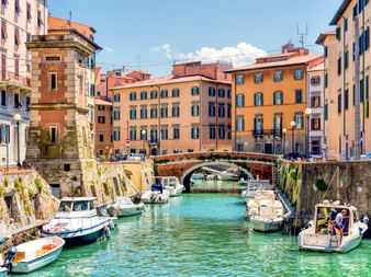 Historic canal in Livorno with moored boats, colorful buildings, and a stone bridge. Clear turquoise water reflects the blue sky.