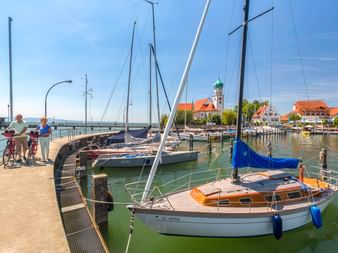 Zwei Radfahrer mit Rädern auf Hafenpromenade in Wasserburg am Bodensee. Segelboote im Hafen, Kirche mit grüner Zwiebelkuppel sichtbar.
