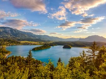 Luftaufnahme des Faaker Sees mit zwei kleinen Inseln, umgeben von Wäldern und Bergen unter dramatischem Sonnenuntergangshimmel.