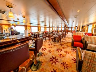 Innenraum der MS Rigoletto mit geräumiger Lounge, Bar, Esstischen, gepolsterten Stühlen, Blumenteppich und großen Fenstern an beiden Seiten.