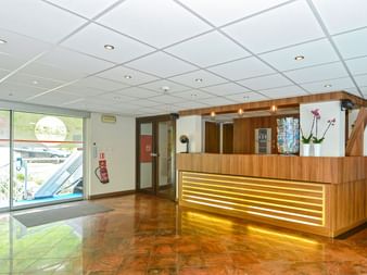 Moderne Rezeption auf der MS Normandie mit beleuchtetem Holztresen, weißer Orchidee und poliertem braunem Boden. Große Fenster zeigen Blick aufs Deck.