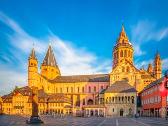 Mainzer Dom mit sechs Türmen in goldenem Sonnenlicht auf einem Stadtplatz in Hessen, Deutschland. Blauer Himmel mit zarten Wolken darüber.