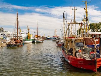 Rotes Fischerboot namens Yvonne im Hafen von Büsum mit anderen Schiffen und weißen Gebäuden unter blauem Himmel mit Wolken.