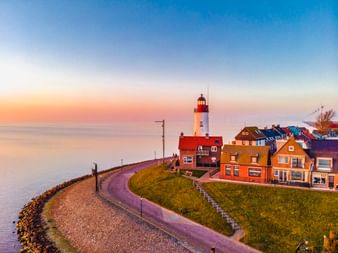 Weißer Leuchtturm mit roter Spitze in Urk bei Sonnenuntergang, umgeben von bunten niederländischen Häusern entlang eines Deichwegs.