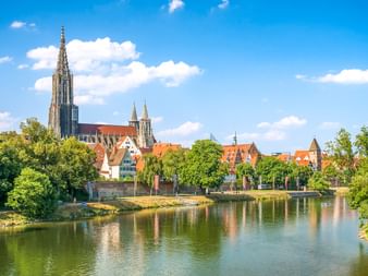 Ulmer Münster mit hohem Turm überragt historische Gebäude mit orangen Dächern entlang der Donau. Grüne Bäume säumen das Flussufer unter blauem Himmel.