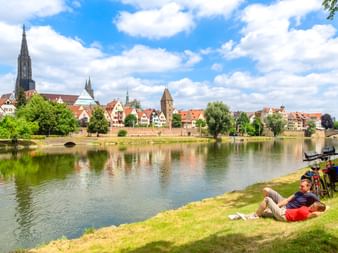 Zwei Radfahrer ruhen auf der Wiese an der Donau in Ulm. Historische Skyline mit Ulmer Münster und bunten Gebäuden spiegelt sich im Wasser.