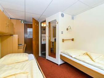 Kompakte Kabine auf der MS Normandie mit zwei Einzelbetten, Holzmöbeln, rotem Teppich und offener Tür zum Bad mit Waschbecken.