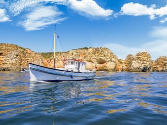 Blau-weißes Fischerboot auf ruhigem Wasser vor goldenen Sandsteinklippen der Algarve-Küste bei Albufeira unter blauem Himmel mit Wolken.