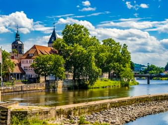Blick auf Hamelns Flussufer mit historischen Gebäuden, Kirchtürmen und grünen Bäumen entlang der Weser unter blauem Himmel mit weißen Wolken.