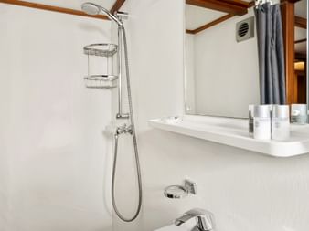 Kompaktes Badezimmer auf der Floor van Aemstel mit weißem Waschbecken, Handbrause, Spiegel und Holzdeckenbalken. Weiße Wände mit Chromarmaturen.