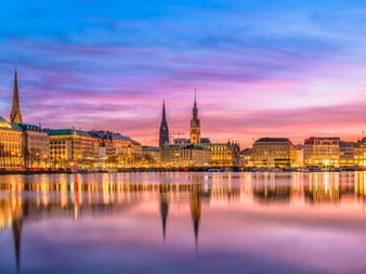 Hamburger Binnenalster bei Sonnenuntergang mit beleuchteten Gebäuden und Kirchtürmen, die sich im ruhigen Wasser unter rosa-blauem Himmel spiegeln.