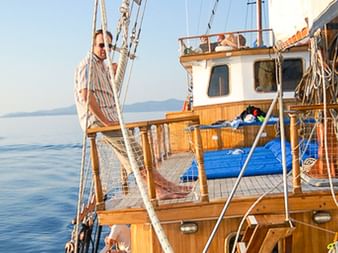 Holzdeck der MS Panagiota mit Geländer und Takelage. Eine Person steht an Deck, ruhige See und ferne Berge im Hintergrund sichtbar.