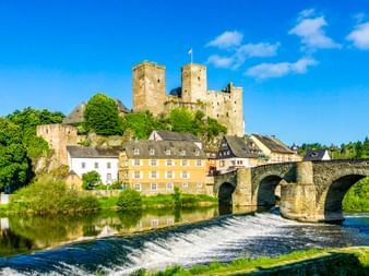 Schloss Runkel thront über einer mittelalterlichen Steinbrücke über die Lahn mit Wehr. Bunte Häuser schmiegen sich unter die Burg an einem sonnigen Tag.