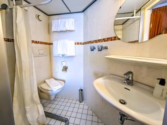 Kompaktes Badezimmer auf der MS Rigoletto mit weißen Fliesen, Waschbecken, Toilette, Duschvorhang, Spiegel und Mosaikbordüre.