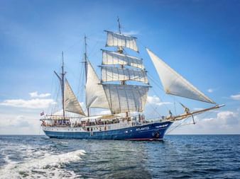 Blauer Großsegler Atlantis segelt mit voll gesetzten weißen Segeln unter blauem Himmel mit vereinzelten Wolken auf offenem Wasser.
