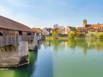 Hölzerne Holzbrücke über den türkisfarbenen Rhein in Bad Säckingen mit historischen Stadtgebäuden und Kirchturm im Hintergrund unter blauem Himmel.