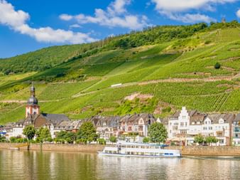 Zell an der Mosel am Moselufer mit Ausflugsschiff, Kirchturm und terrassierten Weinbergen an steilen Hängen unter blauem Himmel.