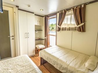 Helle Zweibettkabine auf der MS Olmissum mit zwei Betten, cremefarbenen Wänden, Holzboden, Schrank, Waschtisch und Fenster mit braunen Vorhängen.
