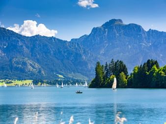 Forggensee bei Füssen mit Segelbooten auf ruhigem blauen Wasser, bewaldeter Halbinsel und dramatischen Berggipfeln unter blauem Himmel.