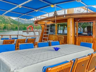 Außenessbereich auf MS Thalassa Deck mit Holztischen, blauen Stühlen unter blauem Sonnensegel. Holzkabine mit Fenstern hinter dem Essbereich.