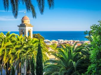 Glockenturm umrahmt von Palmen mit Blick auf die Küste von Sanremo und das blaue Mittelmeer, mit roten Ziegeldächern im Vordergrund.