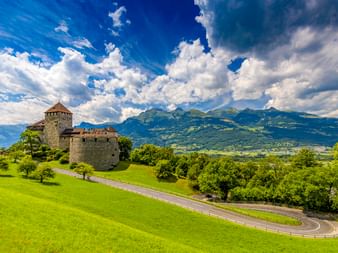 Schloss Vaduz auf einem grünen Hügel mit kurvenreicher Straße, umgeben von Bäumen. Berge und dramatische Wolken im Hintergrund.