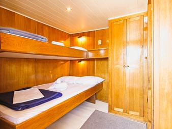 Kompakte Kabine auf der MS Sveti Vid mit Holz-Etagenbetten, weißer Bettwäsche, blauen Handtüchern, Kleiderschrank und warmer Holzverkleidung.