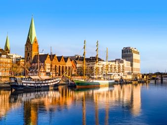 Bremer Hafen mit Segelschiffen am Ufer. Historische Kirche mit grünem Turm und traditionelle Gebäude spiegeln sich im ruhigen Wasser.