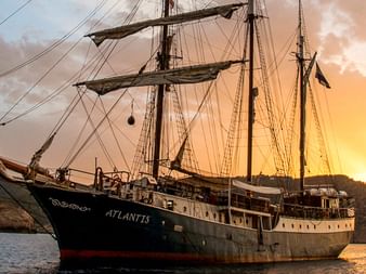 Dreimaster-Segelschiff Atlantis vor Anker in einer Bucht nahe Felsklippen bei Sonnenuntergang mit orangefarbenem Himmel.