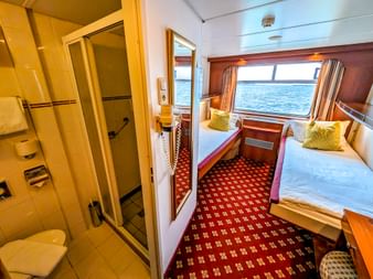 Innenansicht einer MS Rigoletto Kabine mit Einzelbett, gelben Kissen, großem Fenster mit Meerblick, rot gemustertem Teppich und eigenem Bad mit Dusche.