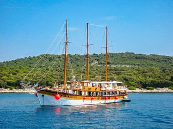 Dreimastiges Holzschiff MS Sveti Vid mit roten Fendern vor Anker in ruhigem blauen Wasser. Grüne bewaldete Hügel im Hintergrund unter klarem Himmel.