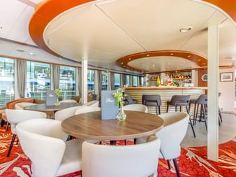 Moderne Lounge auf der MS Lisabelle mit weißen geschwungenen Stühlen, runden Holztischen und einer Bar mit orangefarbenem Deckendesign.