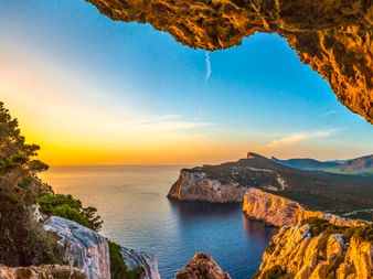 Blick aus einer Höhlenöffnung am Capo Caccia auf dramatische Kalksteinklippen im Mittelmeer bei Sonnenuntergang mit goldenem Licht.