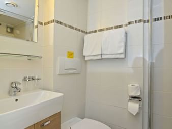 Kompaktes Badezimmer auf der MS Normandie mit weißen Fliesen, Mosaikbordüre, Waschbecken mit Holzunterschrank, WC, Glasdusche und weißen Handtüchern.