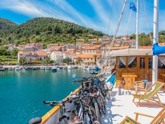 Schiffsdeck mit Fahrrädern entlang der Reling und Liegestühlen, mit Blick auf den Hafen von Korčula mit Häusern und Hügeln.