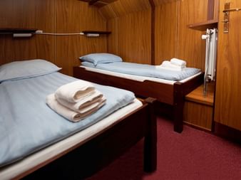 Kompakte Zweibettkabine an Bord der MS Vita Pugna mit zwei Einzelbetten, hellblauer Bettwäsche, gefalteten Handtüchern, Holzverkleidung und rotem Teppich.