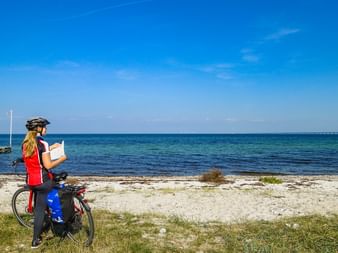 Radfahrerin mit Helm liest Karte am Sandstrand in Dänemark. Türkisfarbenes Meer und blauer Himmel im Hintergrund, Brücke am Horizont.