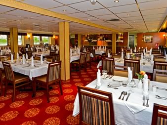 Elegantes Restaurant an Bord der MS Normandie mit weiß gedeckten Tischen, Holzstühlen, rot gemustertem Teppich und warmer Beleuchtung.