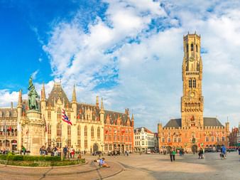 Panoramablick auf den Grote Markt in Brügge mit dem hohen Belfried-Turm, bunten historischen Gebäuden und Besuchern auf dem Platz unter blauem Himmel.
