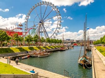 Kiel Germaniahafen harbor with large ferris wheel, historic sailing boats moored in canal, green lawns, and amusement park under blue sky.