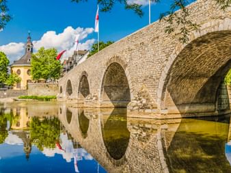 Steinerne Bogenbrücke über die Lahn in Wetzlar mit perfekten Wasserspiegelungen. Kirchturm und Flaggen im Hintergrund unter blauem Himmel.