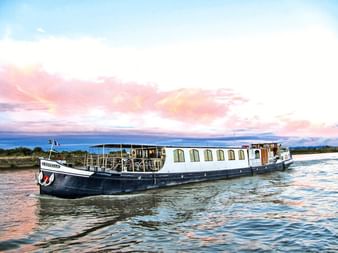 Flusskreuzfahrtschiff MS L'Estello mit weißem Aufbau und dunkelblauem Rumpf auf ruhigem Wasser unter rosa Sonnenuntergangswolken.