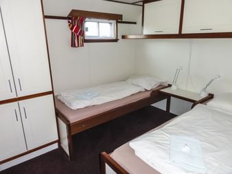 Zweibettkabine auf der MS Lena Maria mit zwei Einzelbetten, weißen Wänden, dunklem Teppich, Einbauschrank und kleinem Fenster über den Betten.