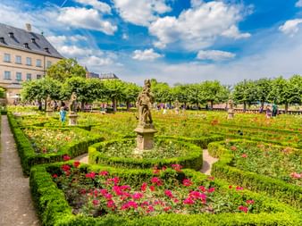 Barocker Rosengarten mit geometrischen Hecken und bunten Rosen an der Neuen Residenz Bamberg. Steinstatue in der Mitte, Palastgebäude dahinter.