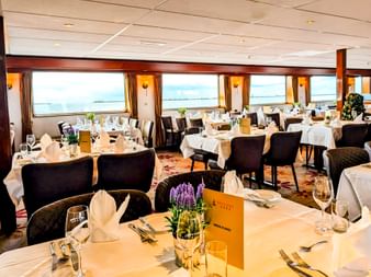 Eleganter Speisesaal an Bord der MS Rigoletto mit weiß gedeckten Tischen, schwarzen Stühlen und großen Fenstern mit Flussblick.