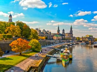 Dresdner Elbufer mit Herbstbäumen, historischer Skyline und Booten. Die barocke Altstadt mit Kirchtürmen ist über den Fluss sichtbar.