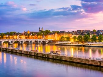 Historische Steinbrücke mit beleuchteten Bögen über die Maas in Maastricht bei Dämmerung. Bunte Gebäude säumen das Ufer unter violettem Himmel.