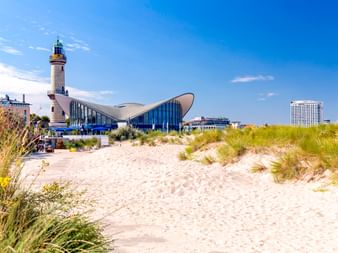 Sandstrand mit Dünengras führt zum Rostocker Teepott und weißem Leuchtturm. Moderne geschwungene Architektur mit blauer Glasfassade unter klarem Himmel.