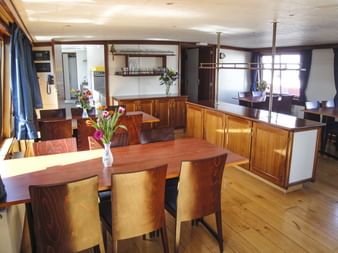 Restaurant an Bord der MS Lena Maria mit Holztischen, Stühlen und Schränken. Frische Blumen in Vasen schmücken die Tische. Tageslicht fällt durch die Fenster.