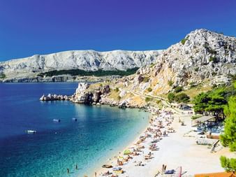 Kieselstrand in Baska, Kroatien mit Badegästen am türkisfarbenen Adriatischen Meer. Weiße Felsengebirge erheben sich dramatisch hinter der Bucht.